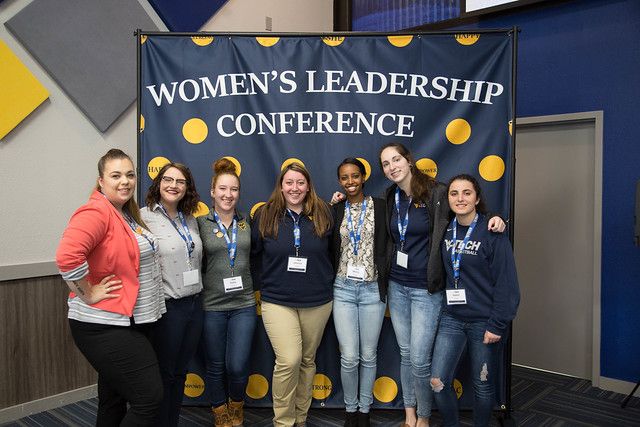 A group of seven women stand for a photo in front of a gold and blue backdrop at the first Women's Leadership Conference