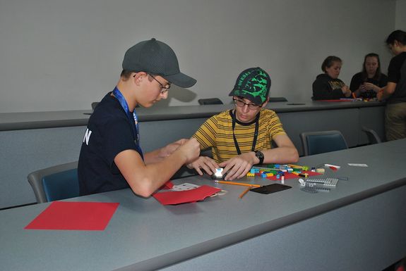 Two young boys with hats on are doing an activity with legos at a long gray table