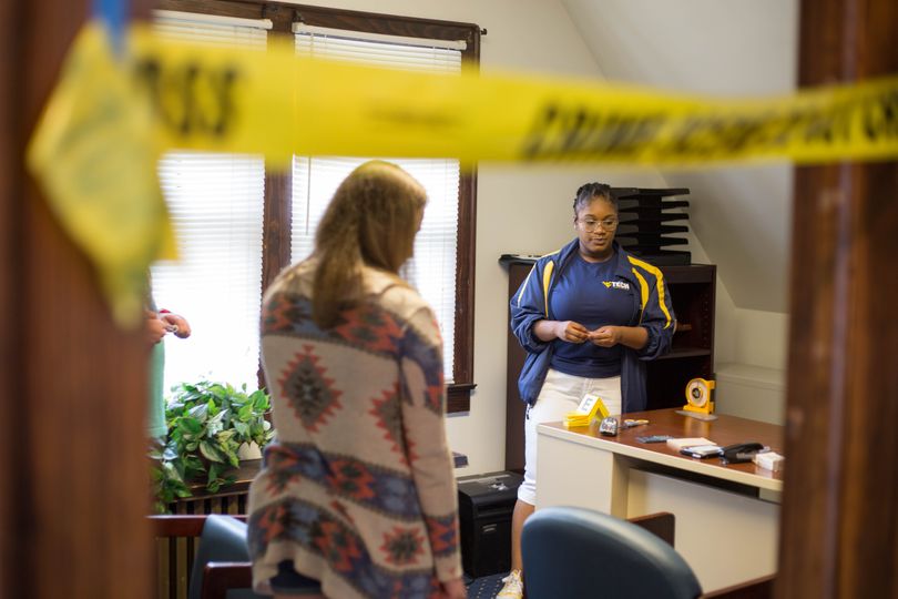 From a first-person perspective, we see a door jamb with crime scene tape running across it. Behind, two investigators stand next to a desk discussing their findings.