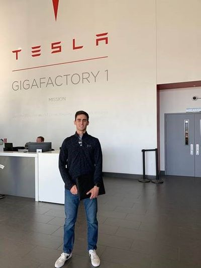 Antonio stands in front of a front desk reception area with the Tesla Gigafactory logo behind him
