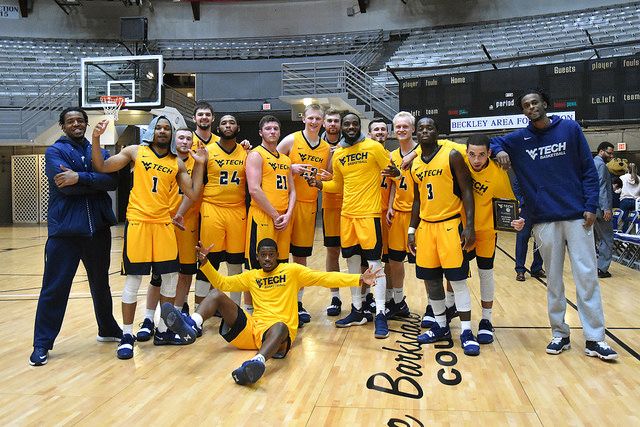 The WVU Tech men's basketball team, dressed in their gold uniforms, stand together alongside coaches for a photo. They're standing on the court. one player is seated and playfully kicking his feet out towards the camera.