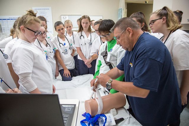A male nurse in blue scrubs shows students dress in their nursing coats how to secure a wound.