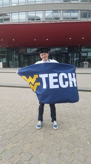 WVU Tech student Cody Walker wears a graduation cap and holds a WVU Tech flag in front of the school in the Netherlands.