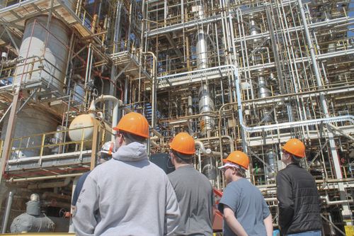 A group of students stand in front of a very intricate looking building with pipes. They are wearing hard hats.