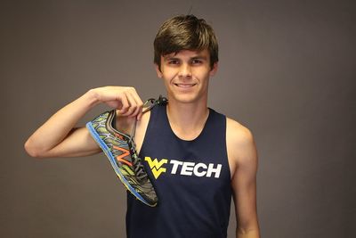 Student Luke Jobson stands in front of a grey background, wearing his cross country jersey and holding a pair of running shoes over his shoulder.