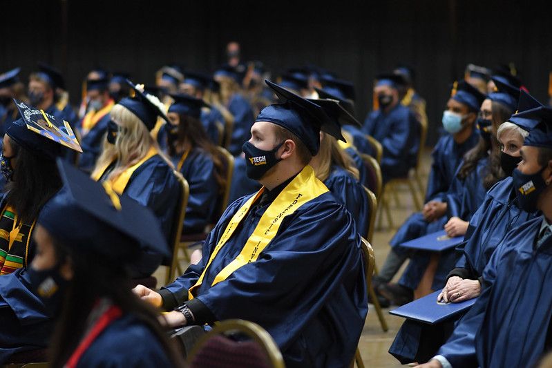 A sea of WVU Tech graduates sit in the convention center, the camera focuses on a male grad in his cap, gown and mask as he listens to the speaker.