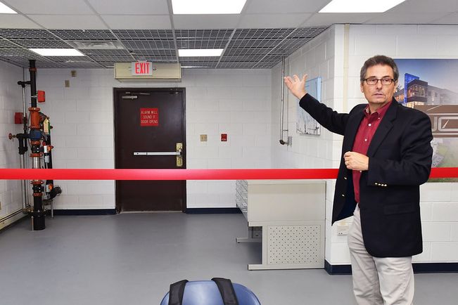 A man in front of a red ribbon gestures behind him, showing the back of the classroom celling that is open. This is for students to be able to see and learn from. 