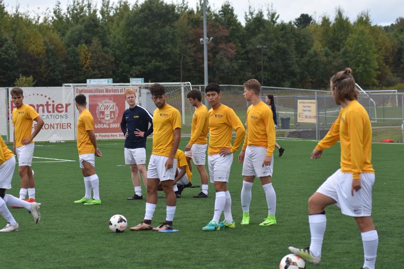 A group of WVU Tech men's soccer players, dressed in gold shirts and white shorts, run drills on a soccer field during the day.