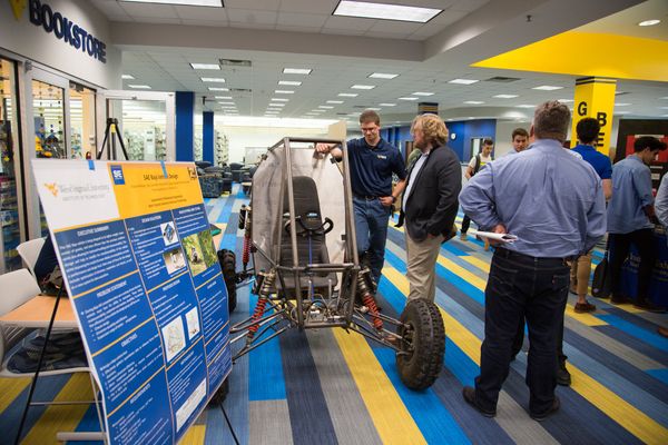A student stands next to a racing vehicle on display. He's motioning to the vehicle as he chats with a man in a suit.