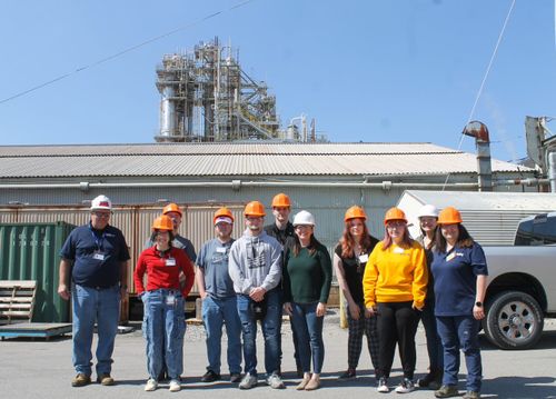 A group of students and professors stand in front of a plain building at the Belle Chemical plant with hard hats on