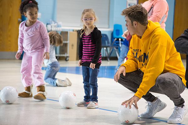 In a gym, a male WVU Tech student crouches to talk to two Head Start students, his hand resting on a small, white soccer ball. To his left, two four-year-old girls listen to him as they wait anxiously to kick the soccer balls at their feet.