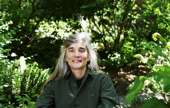 Author and essayist, Ann Pancake, sits in a green shirt, a lush forest behind her, as she looks, smiling, at the camera.