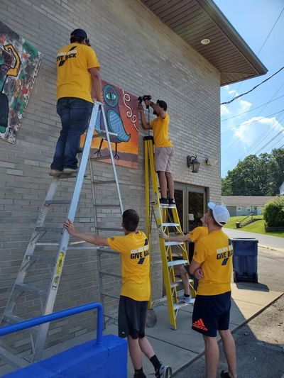 Student volunteers climb a ladder to paint during a service project.