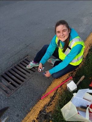 Tech student Katie Stanley sits near a storm drain just off the sidewalk. Her jacket is high-visibility, and so is her smile as she looks up at the camera from her painting work.