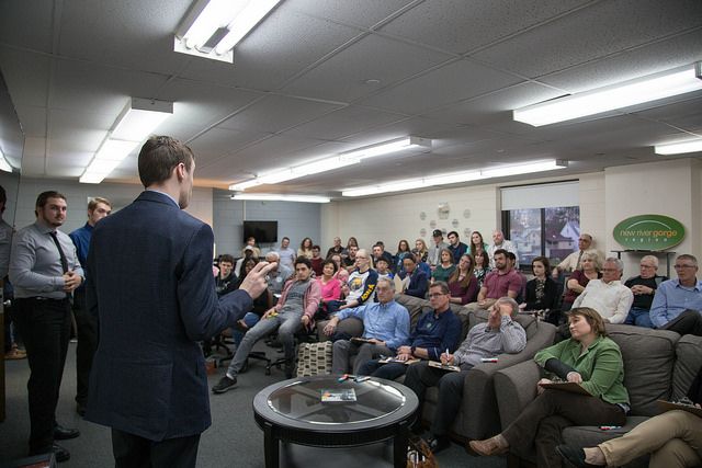 Well-dressed students stand with their backs to the camera, pitching a business idea to a crowd of dozens of people sitting in a conference room.