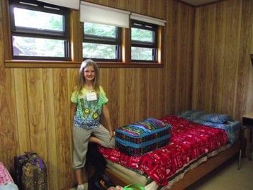 A young girl stands in a cabin like room with a suitcase laying on a bed beside the girl. Alexis Zilinski is wearing a 4H vest.